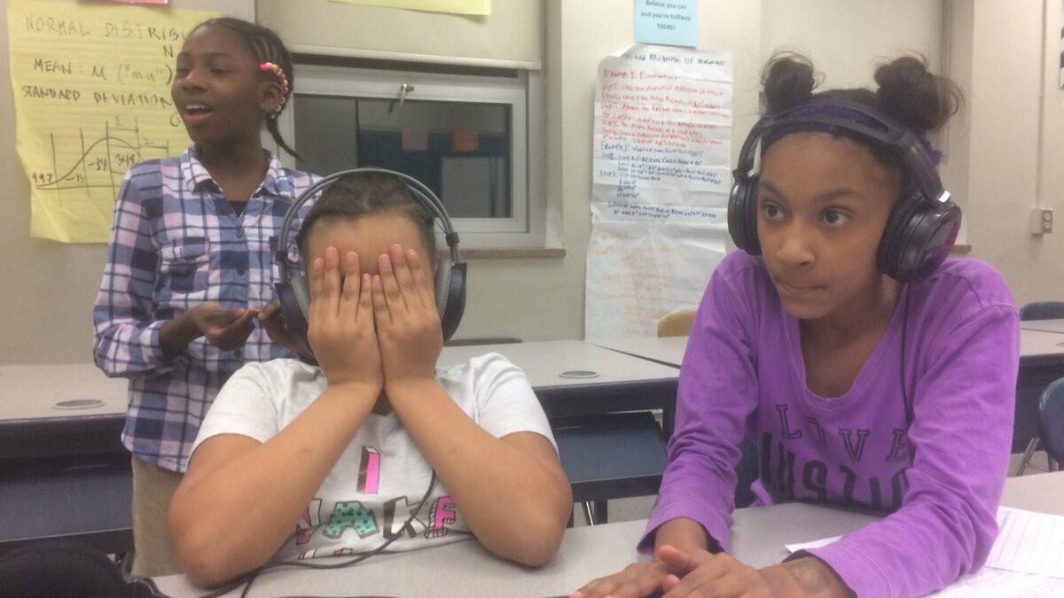 Three youth in a classroom behind a mixer, two wearing headphones, one holding their face in their hands