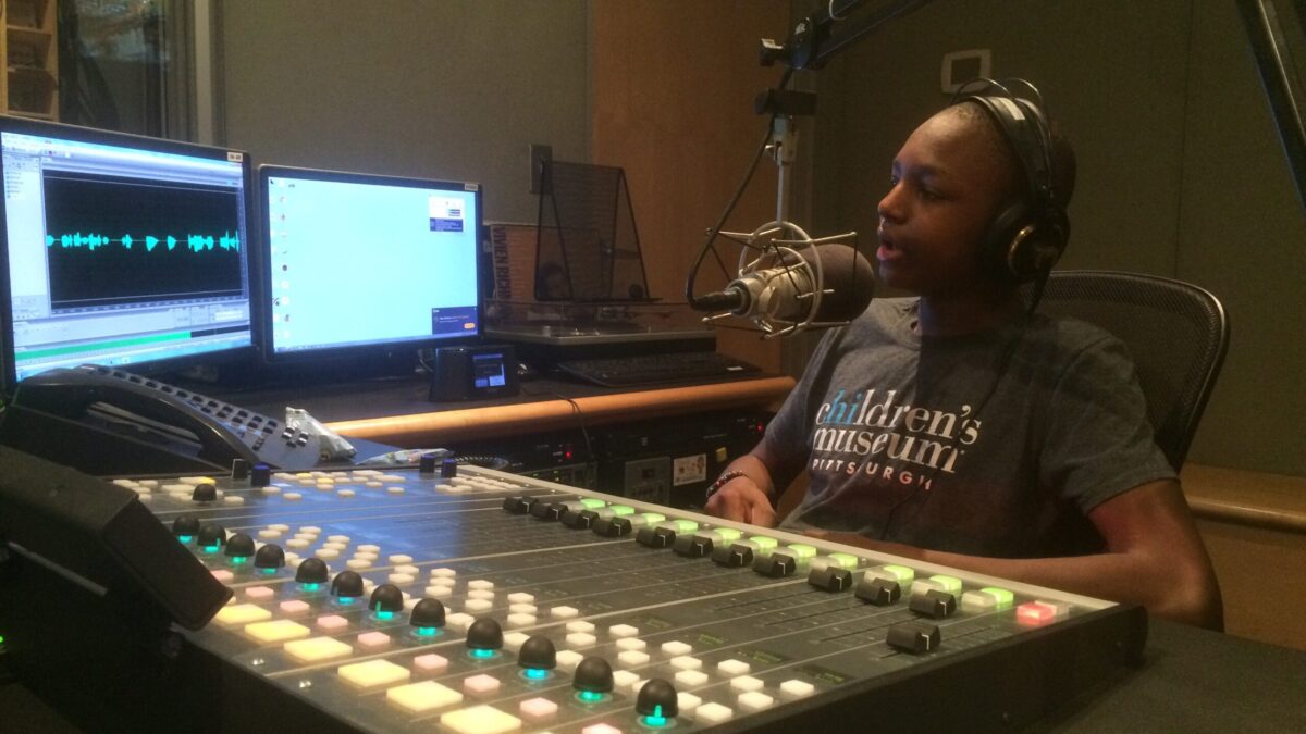 A teen wearing headphones speaking into a microphone sitting behind a mixing board and computer screens in a radio studio