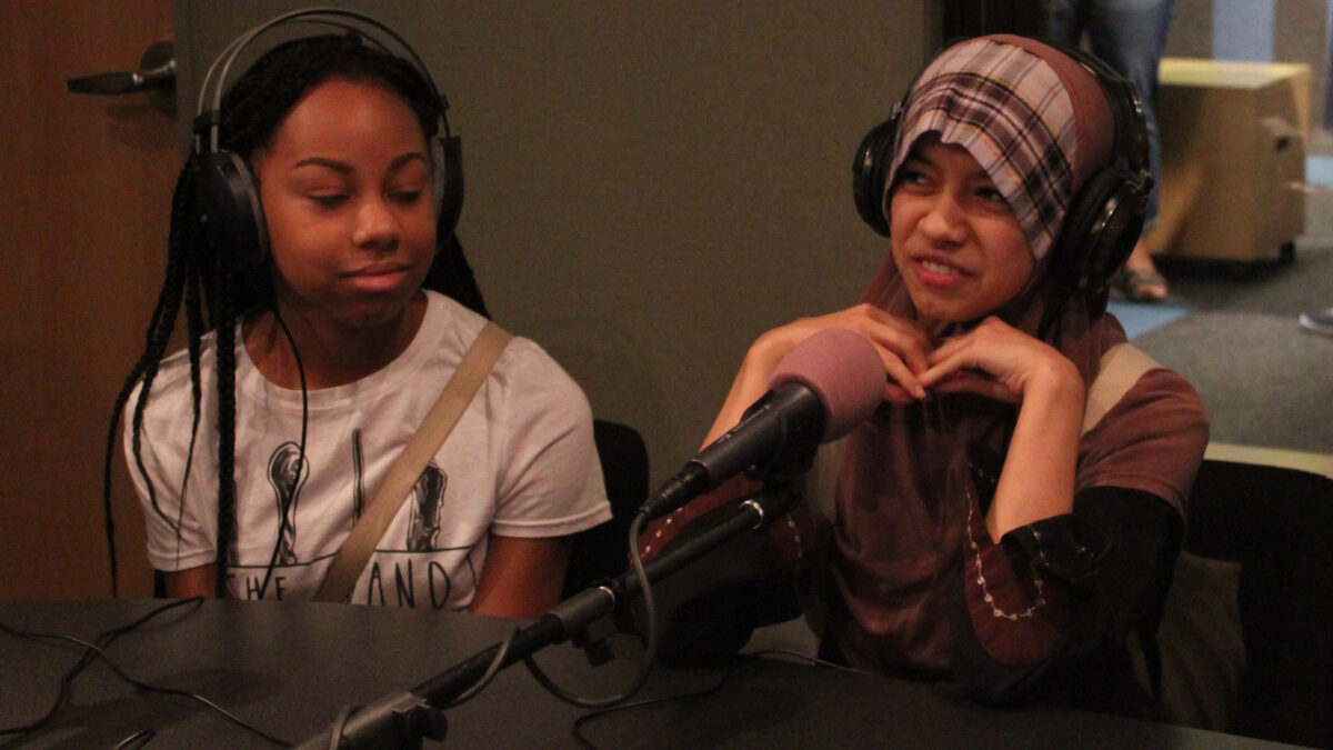 Two teens wearing headphones speaking into microphones in a radio studio