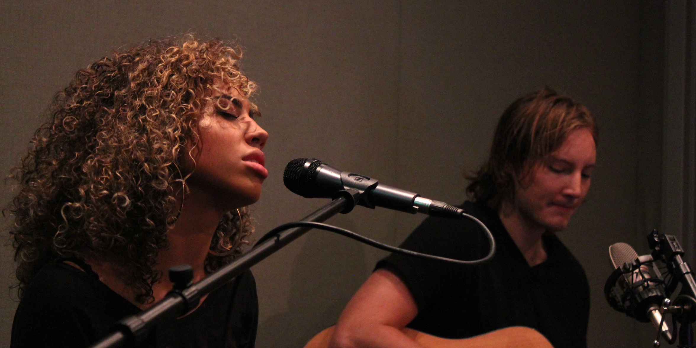 Two adults singing into a microphone, one playing an acoustic guitar, in a radio studio
