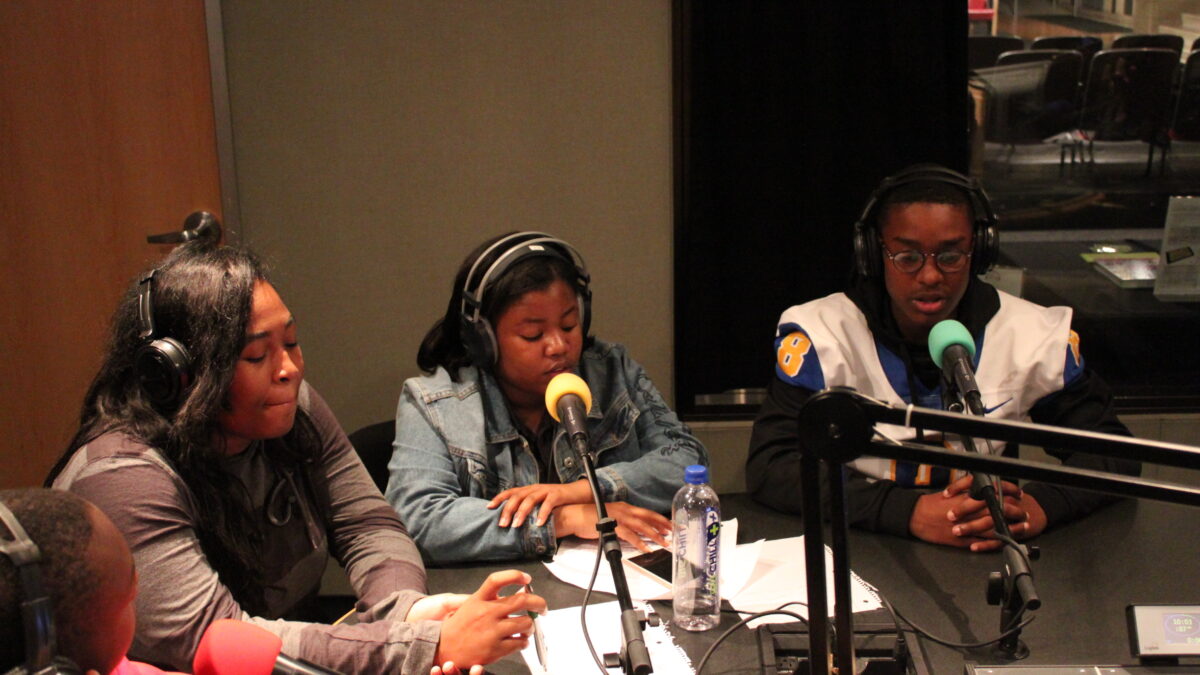 Four teens wearing headphones speaking into microphones in a radio studio