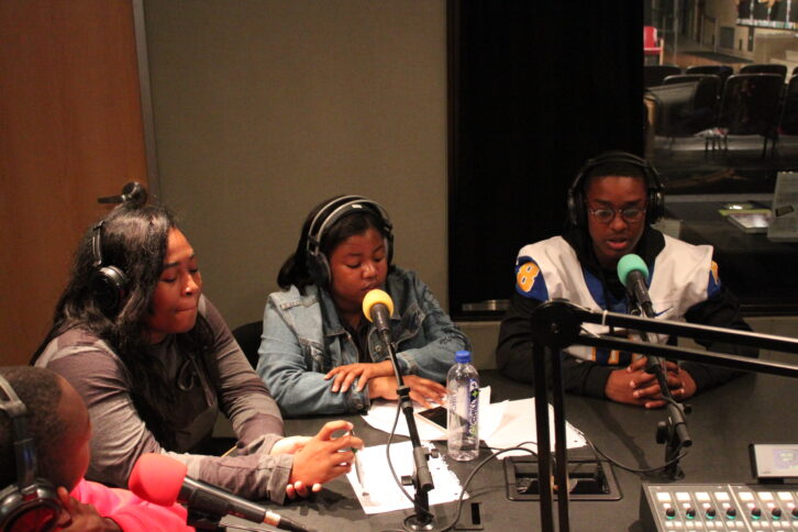 Four teens wearing headphones speaking into microphones in a radio studio