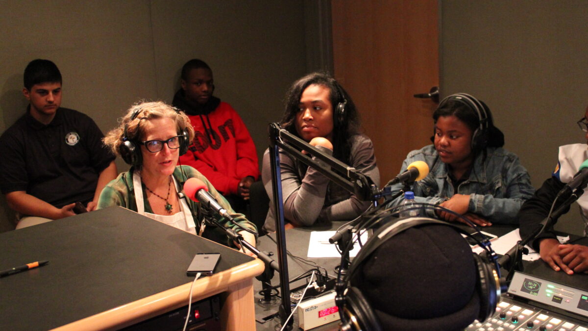Two youth and an adult wearing headphones speaking into microphones as a teen wearing headphones and two teens sitting in the background listen in a radio studio