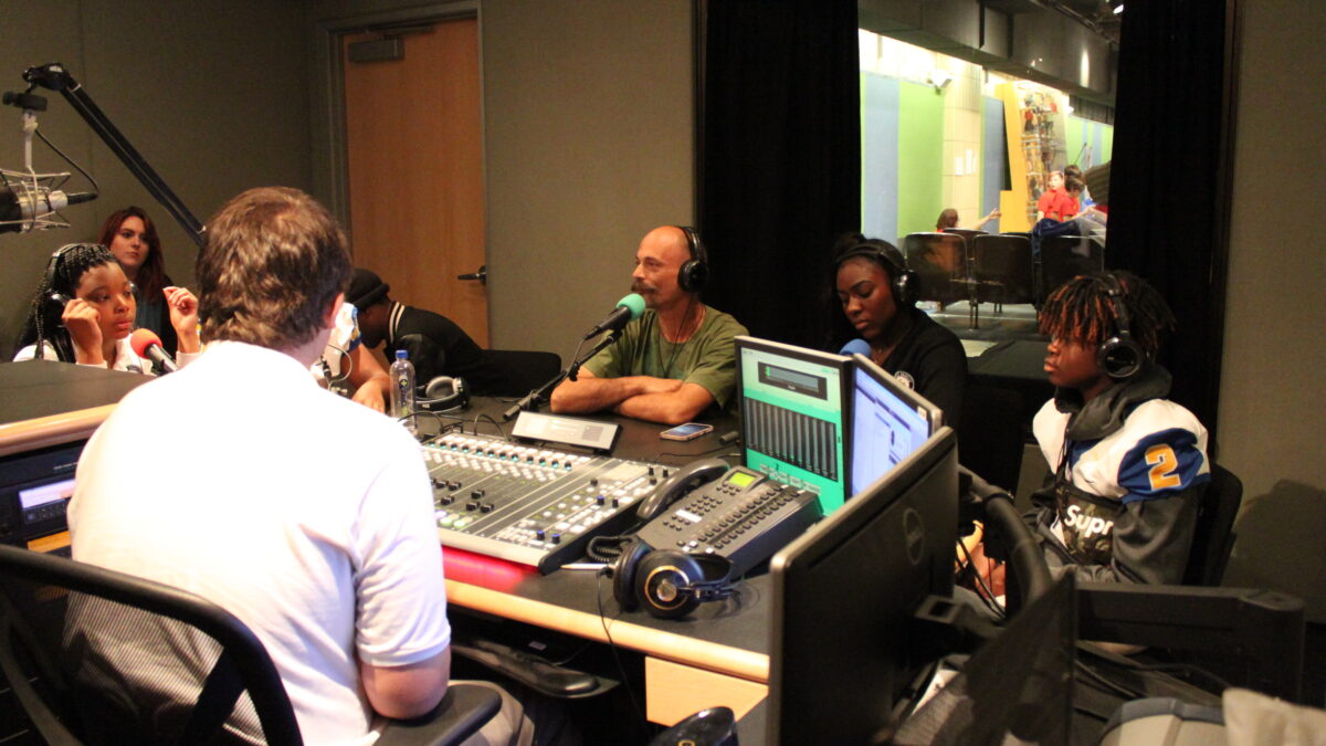 Four teens and an adult wearing headphones speaking into microphones as two adults listen in a radio studio