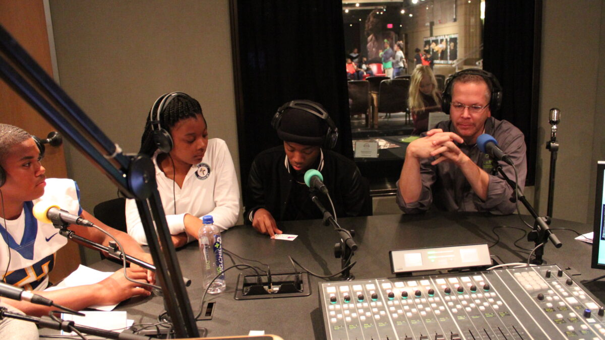Three teens and an adult wearing headphones speaking into microphones in a radio studio