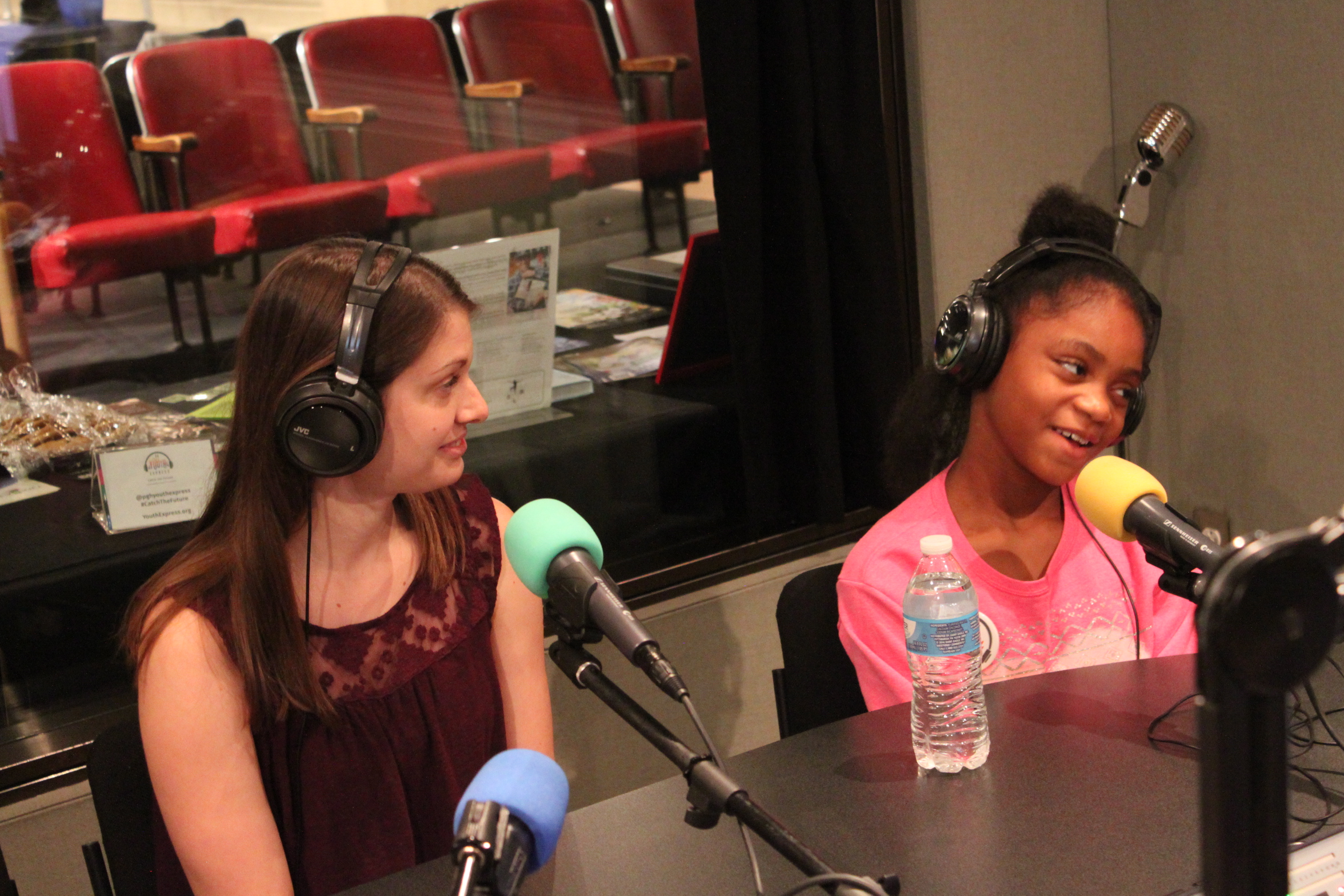 Two individuals wearing headphones speaking into microphones in a radio studio