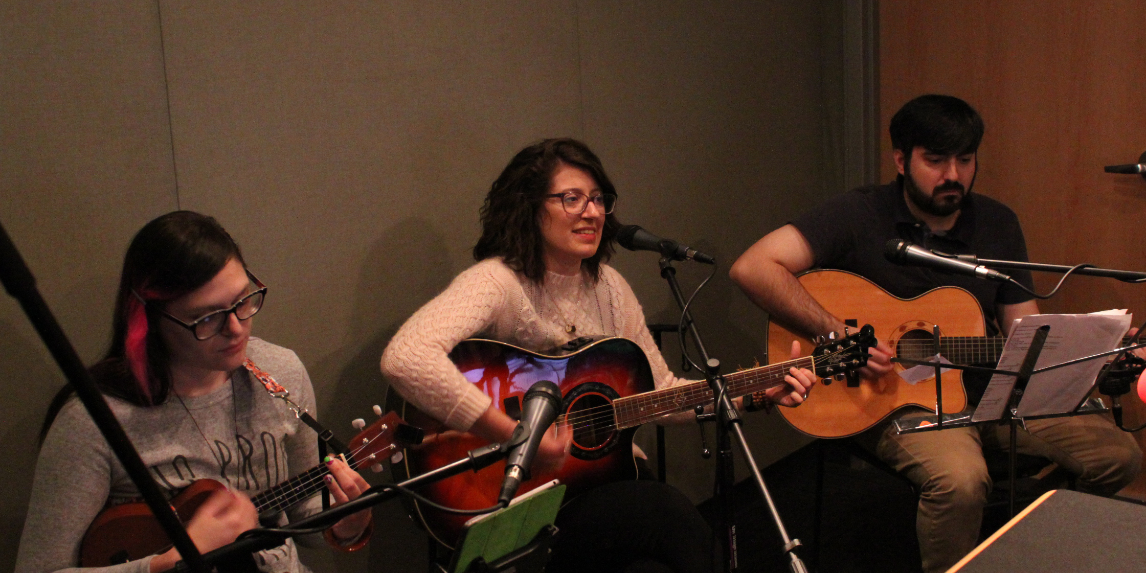 An adult playing acoustic guitar, an adult playing acoustic guitar and singing into a microphone, and an adult playing ukulele in a radio studio