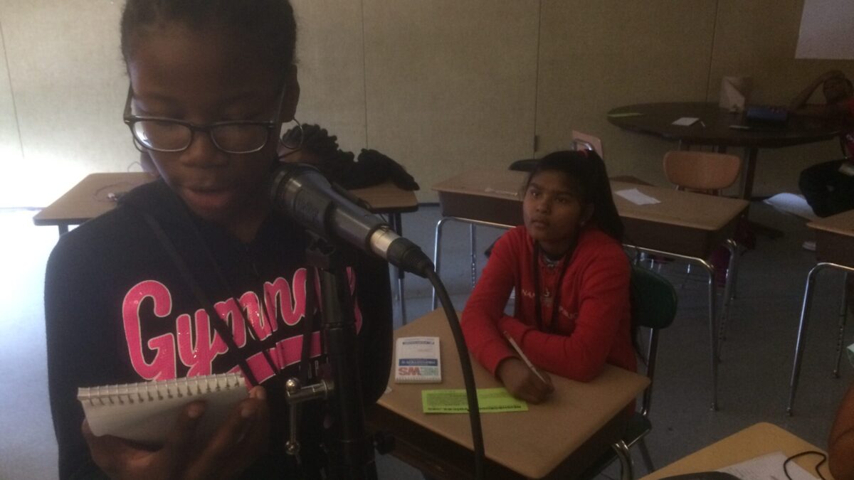 A youth holding a notepad reading into a microphone as another youth listens in a classroom