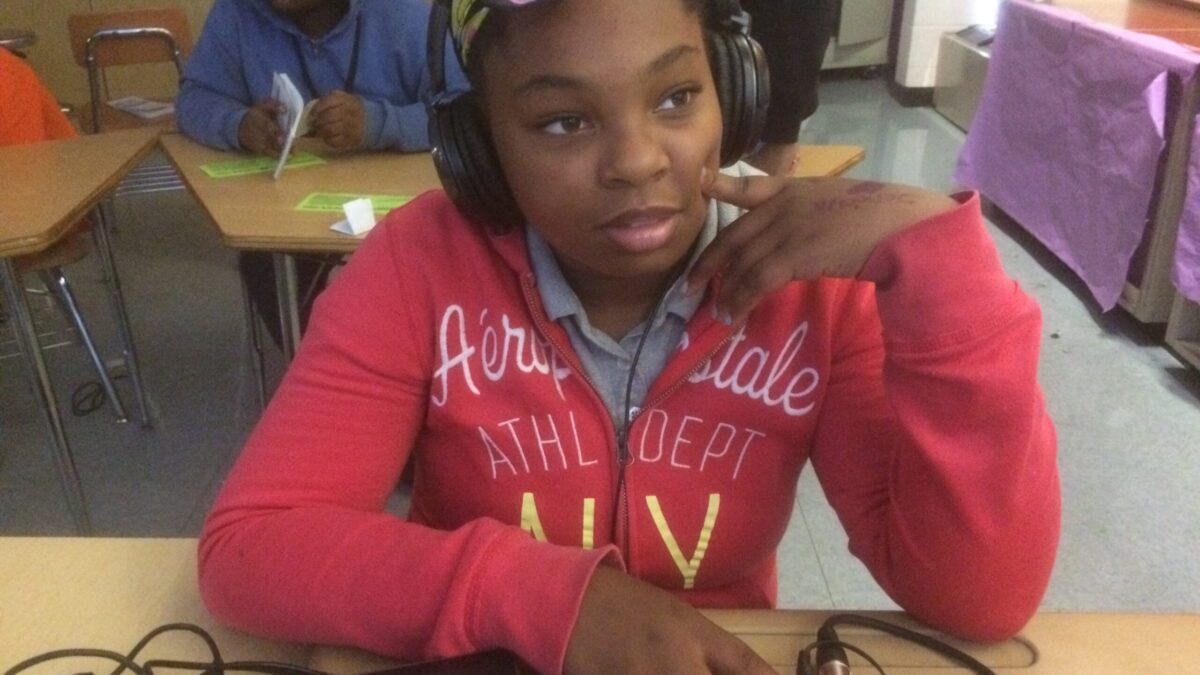A youth wearing headphones sitting at a classroom desk with a portable recorder.