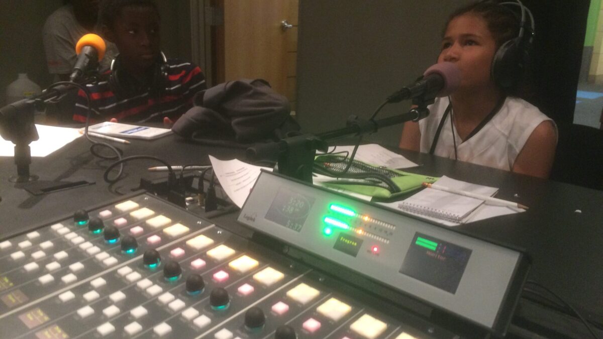 Two youth, one wearing headphones, speaking into microphones in a radio studio