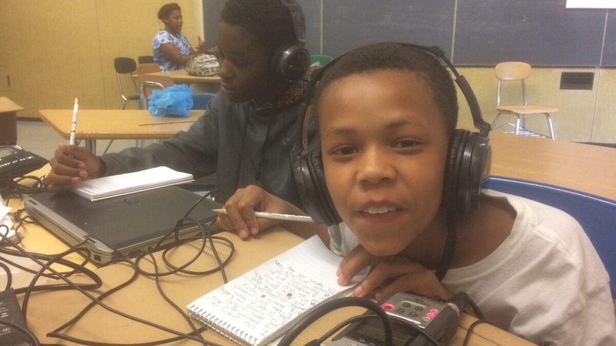 Two youth wearing headphones writing on notepads on a desk in a classroom, one smiling at the camera