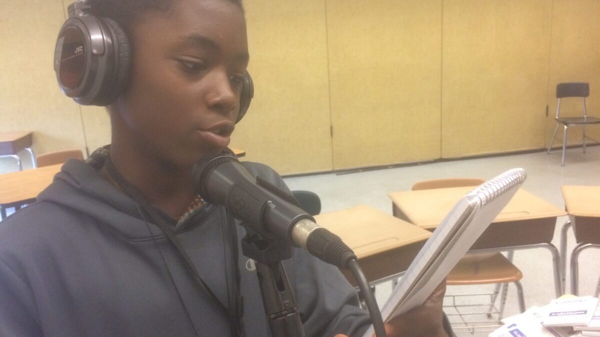 A youth wearing headphones reading from a notepad into a microphone in a classroom.