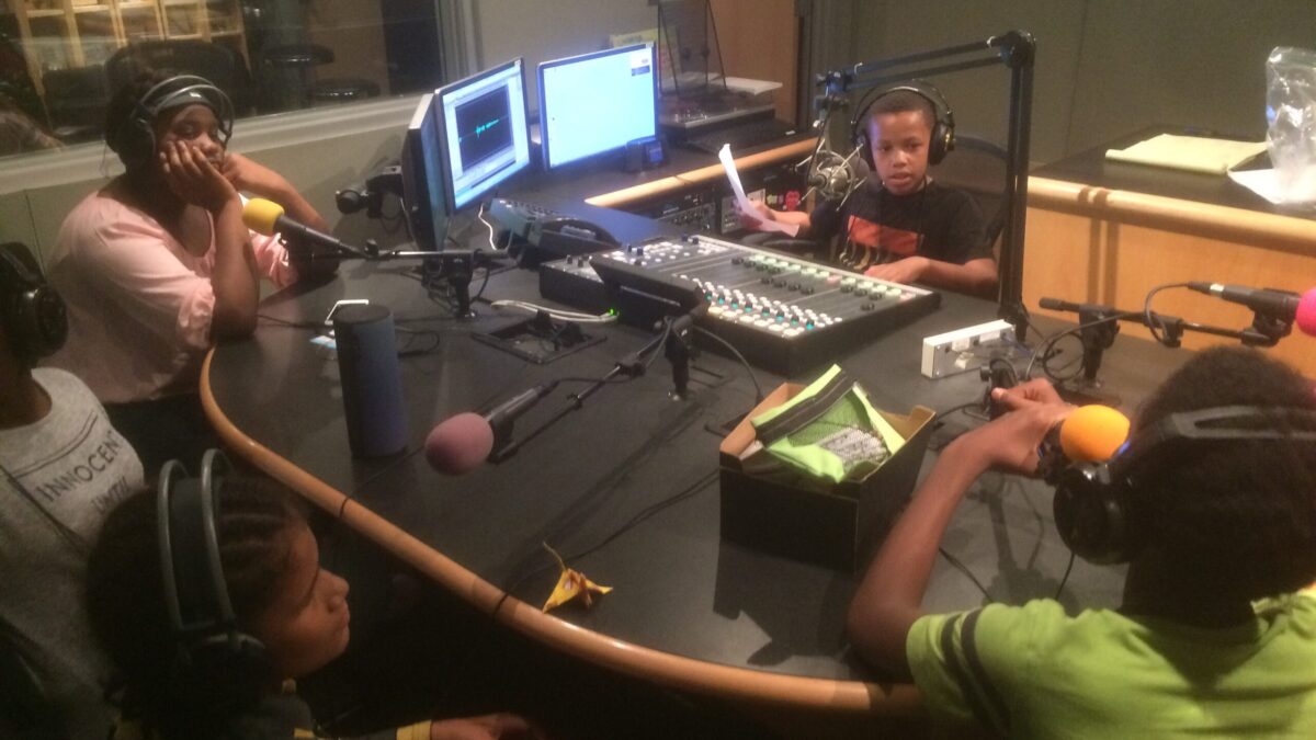 Four youth wearing headphones sitting behind microphones and a youth wearing headphones holding a piece of paper sitting behind a microphone and a mixing board in a radio studio