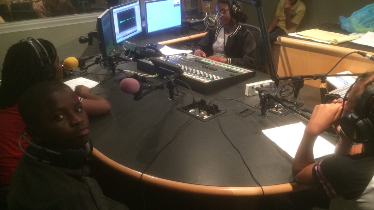 Three youth wearing headphones sitting behind microphones and one youth wearing headphones sitting behind a microphone and a mixing board in a radio studio