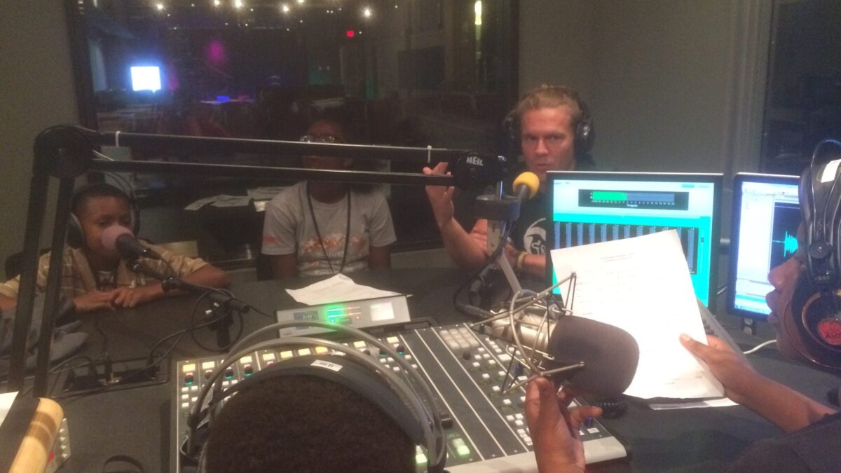 Three youth wearing headphones sitting behind microphones and two youth wearing headphones sitting behind a microphone and a mixing board in a radio studio