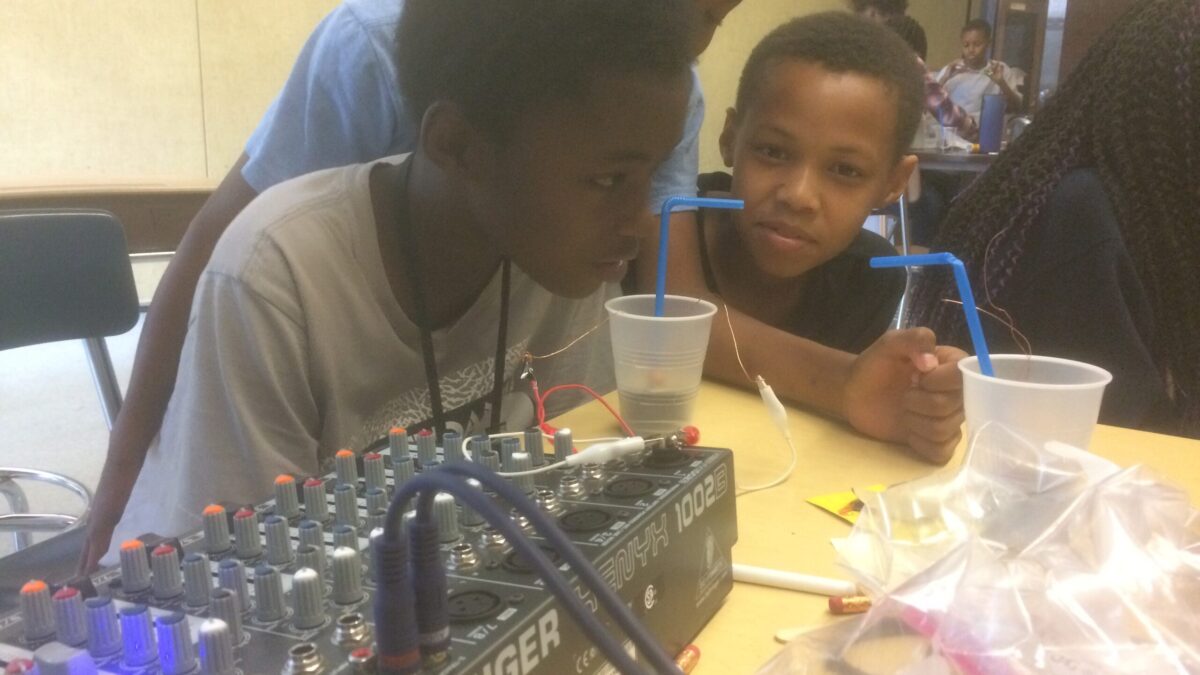 Three youth sitting behind a desk with a mixer and two plastic cups with straw
