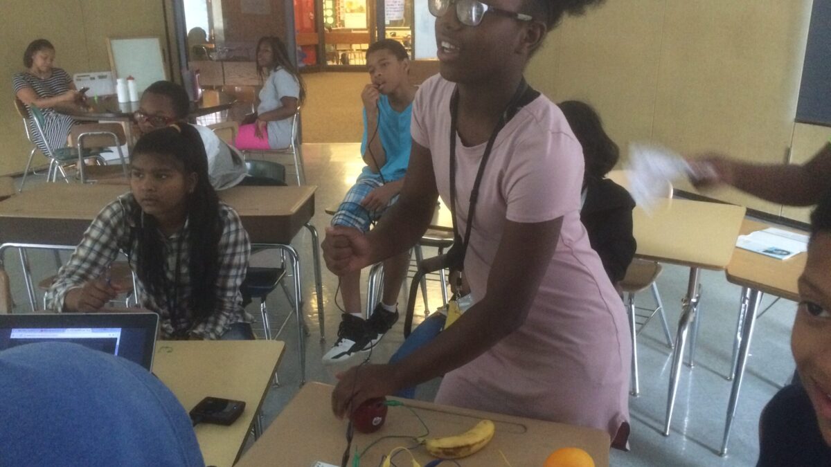 Six youth sitting in a classroom, one standing and gesturing with their hands