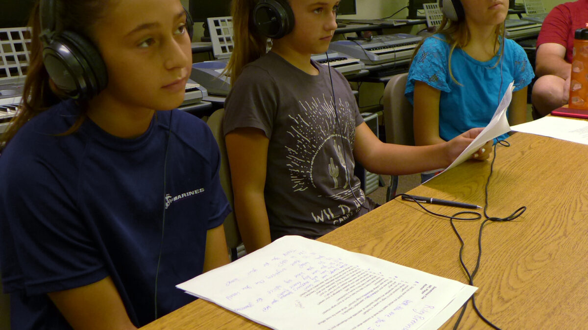 Three teens wearing headphones looking at pieces of paper on a long wooden table