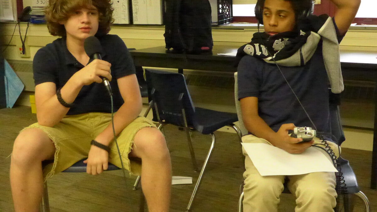 A youth speaking into a microphone and a youth wearing headphones holding a portable recorder in a classroom