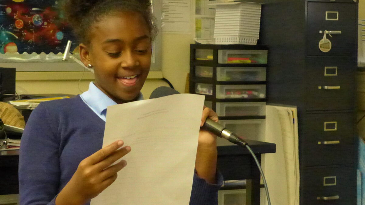 A youth holding a piece of paper speaking into a microphone in a classroom