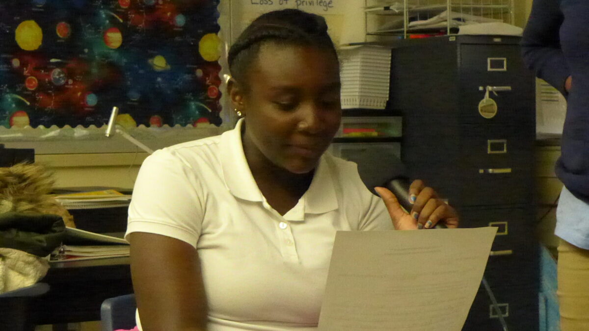 A youth holding a piece of paper and speaking into a microphone in a classroom
