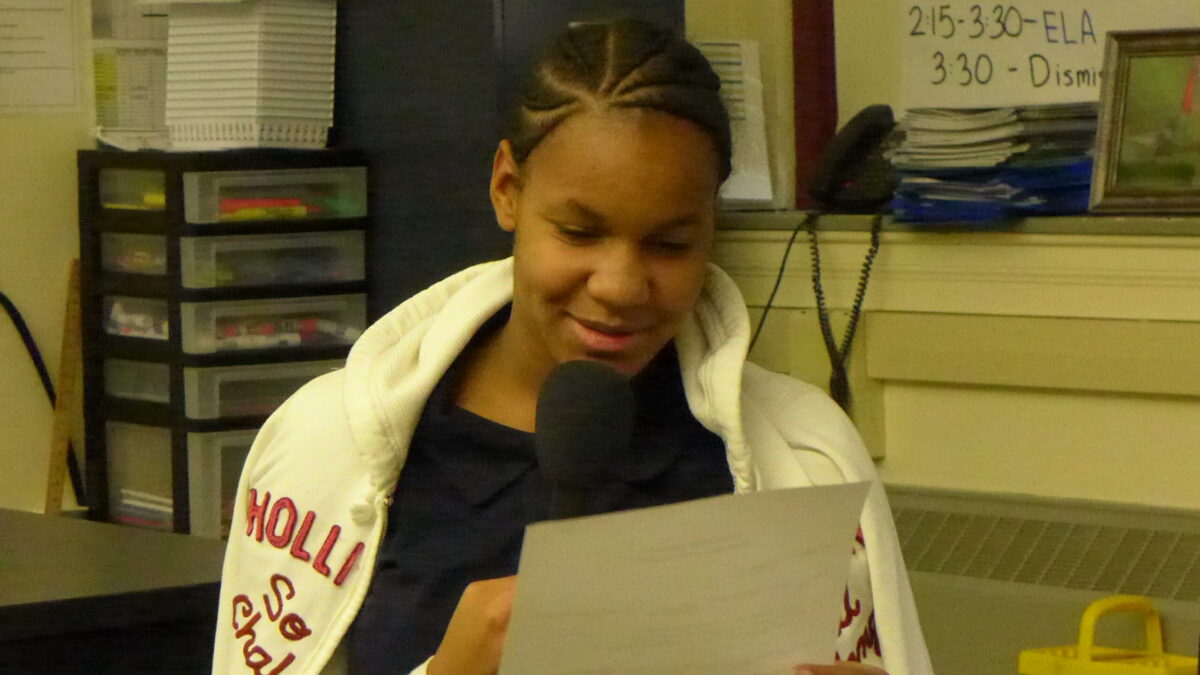 A youth holding a piece of paper speaking into a microphone in a classroom