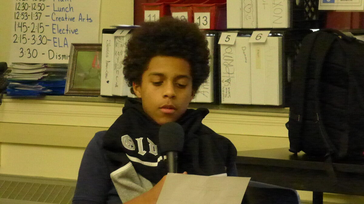 A youth holding a piece of paper speaking into a microphone in a classroom