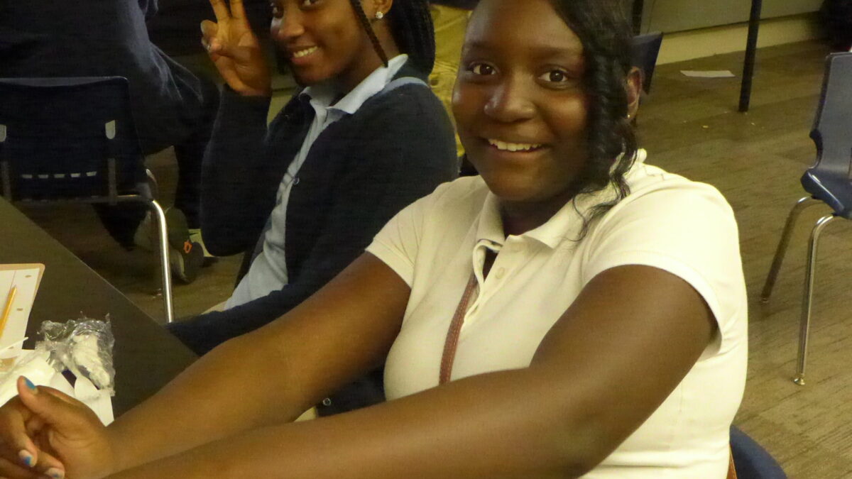 Two youth smiling sitting behind a desk in a classroom, one flashing a peace sign