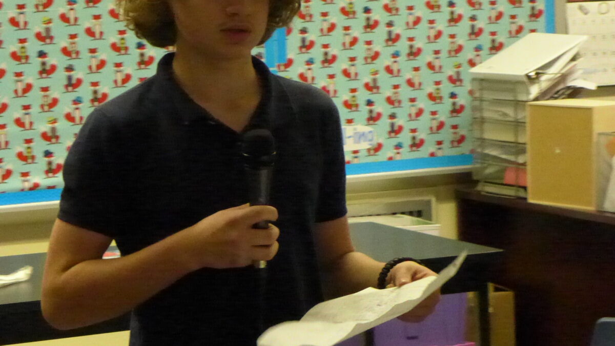 A youth holding a piece of paper speaking into a microphone in a classroom