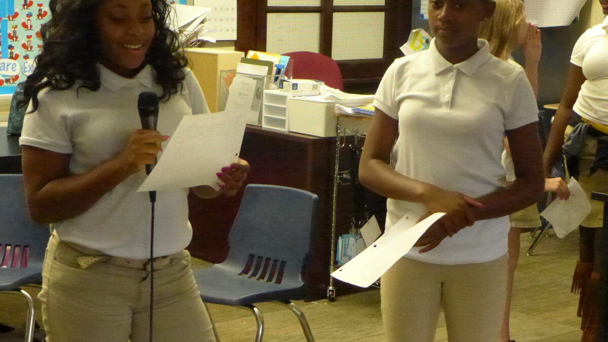 Two youth holding a piece of paper in a classroom, one holding a microphone