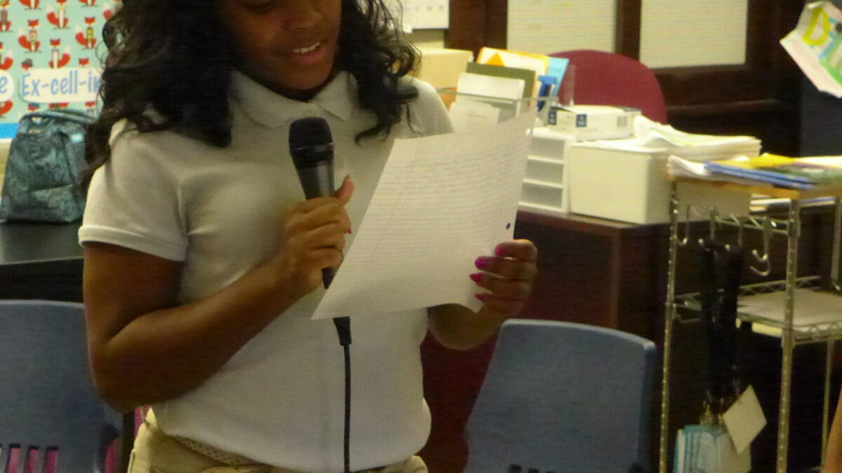 A youth holding a piece of paper speaking into a microphone in a classroom