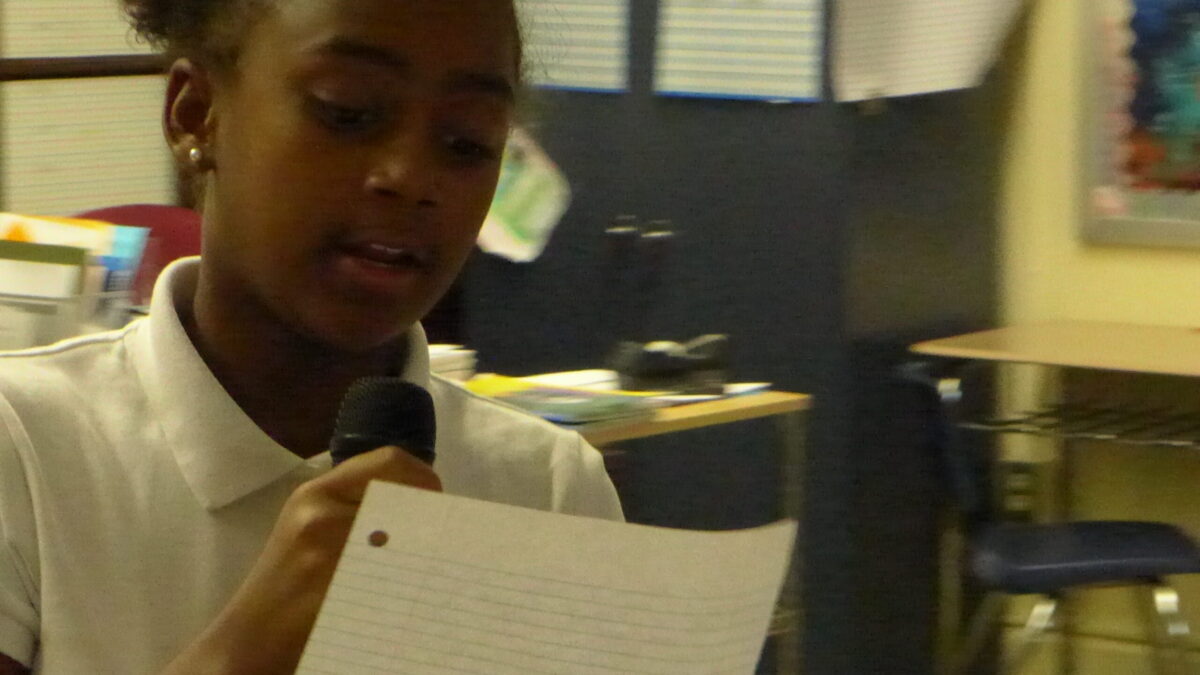 A youth holding a piece of paper speaking into a microphone in a classroom