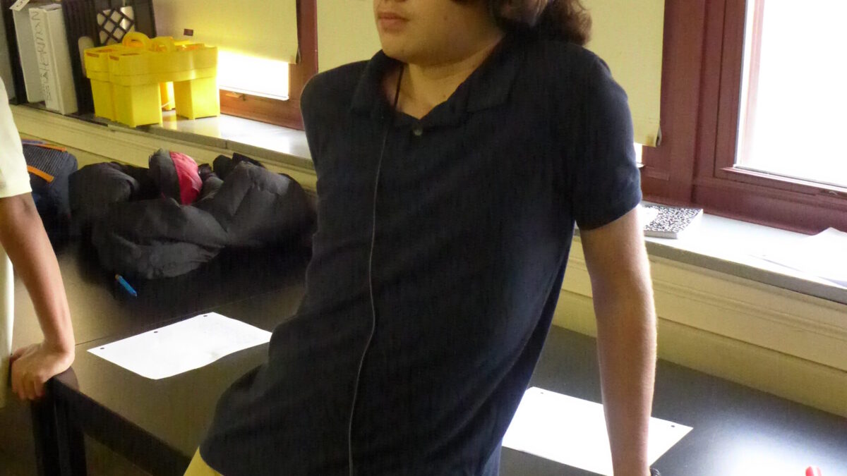 A youth posing on a desk standing next to a portable recorder in a classroom