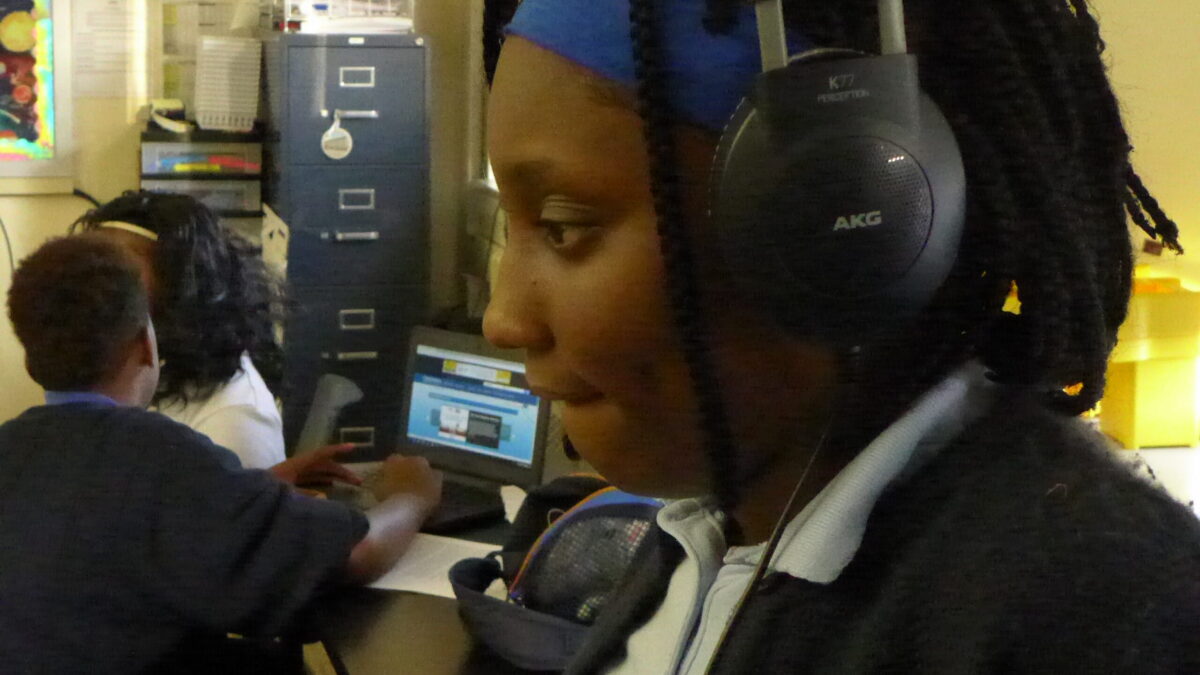 A youth wearing a pair of headphones and an individual interacting with a laptop in a classroom