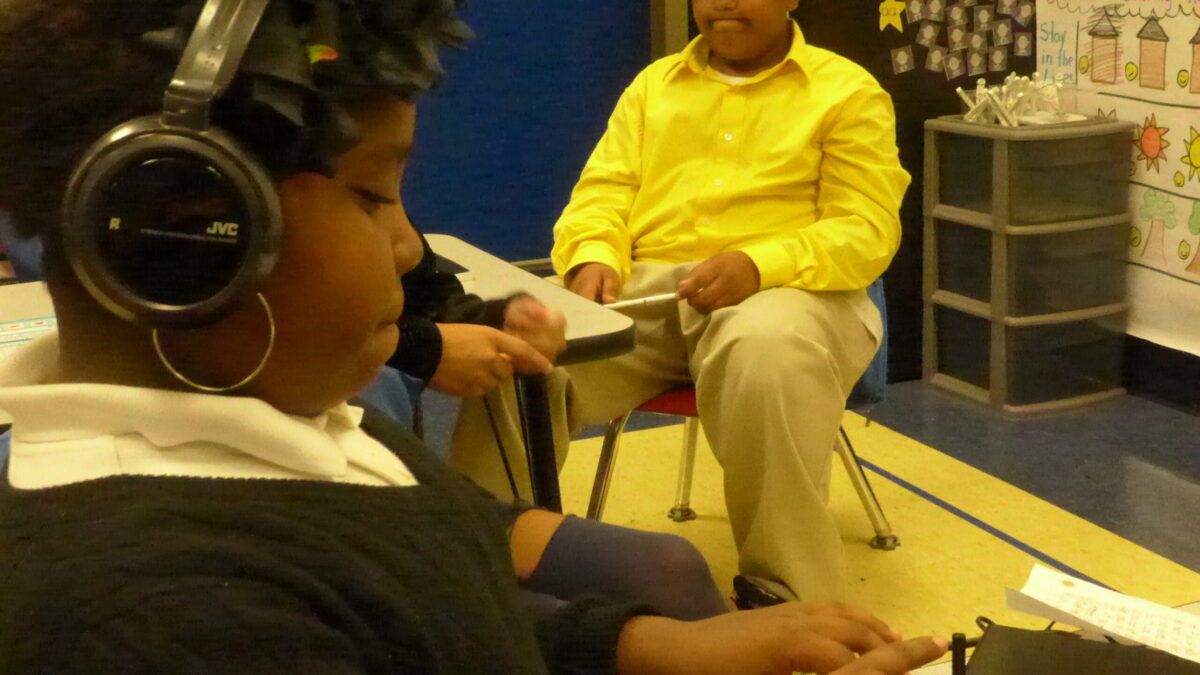 A youth wearing headphones connected to an audio driver and another youth sitting at a desk in a classroom