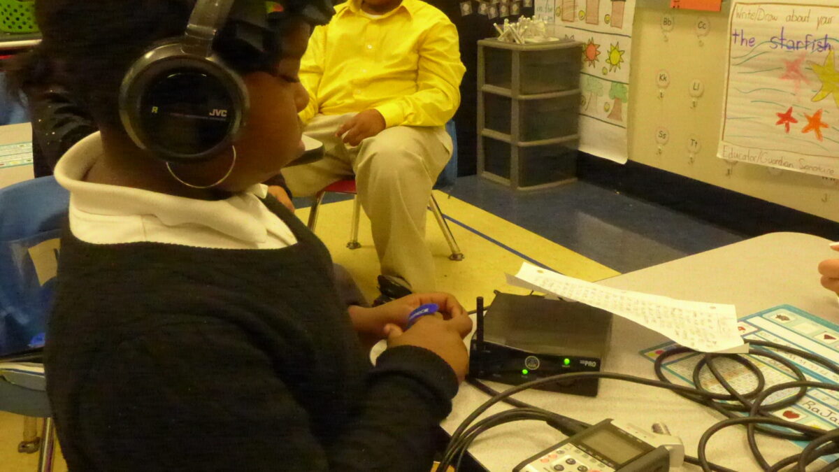 A youth wearing headphones connected to an audio driver and another youth sitting at a desk in a classroom