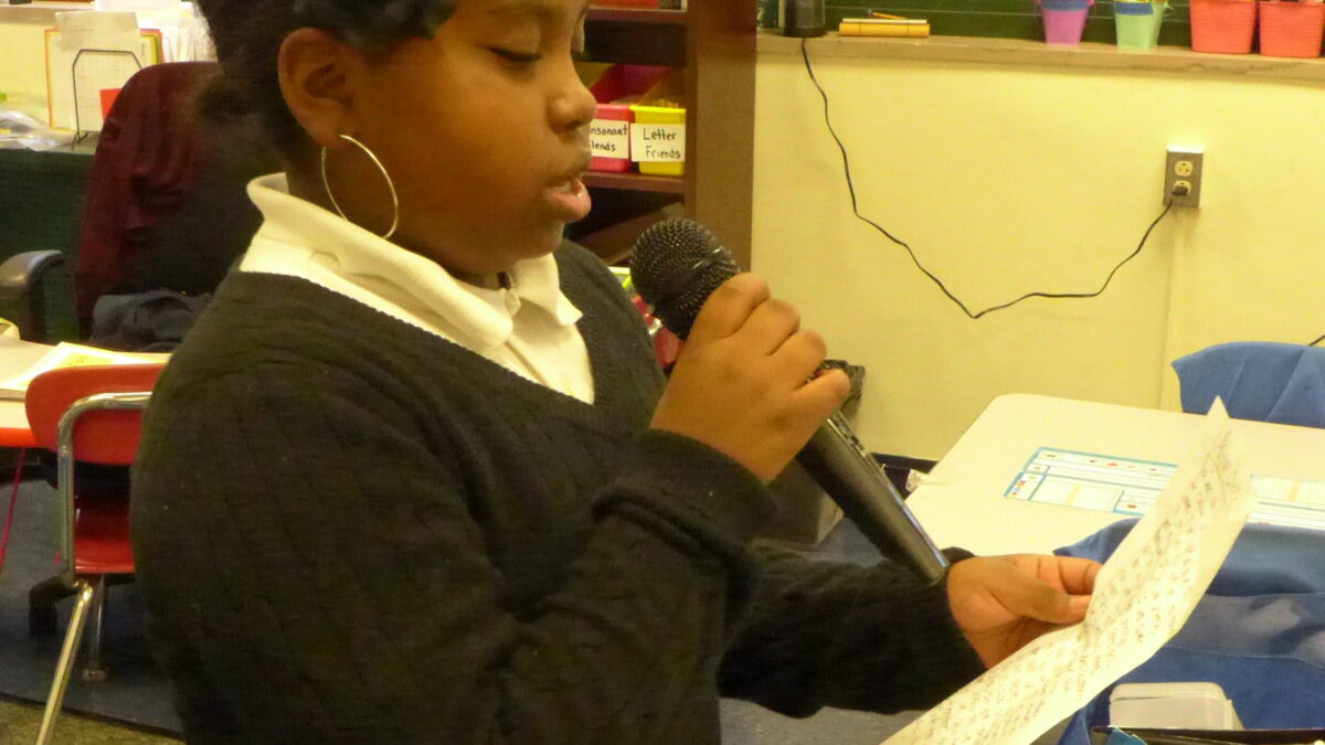 A youth holding a piece of paper speaking into a microphone in a classroom