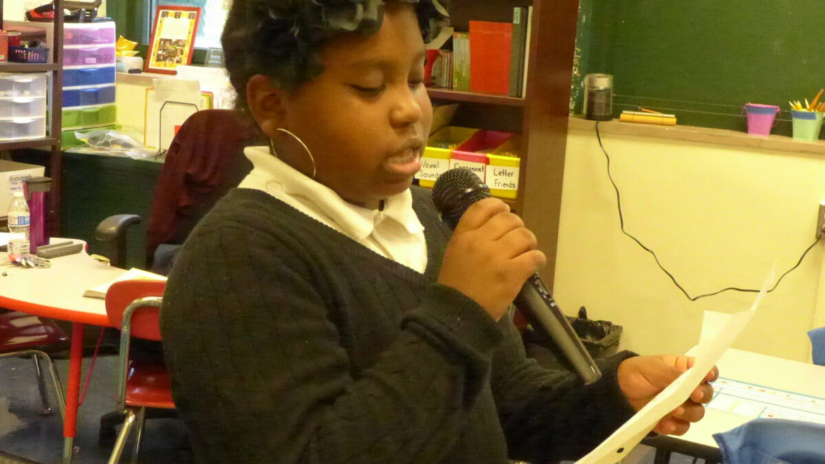A youth holding a piece of paper speaking into a microphone in a classroom