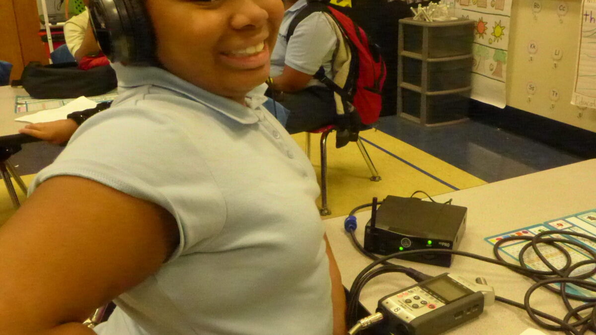 A youth wearing headphones connected to a portable recorder smiling in a classroom
