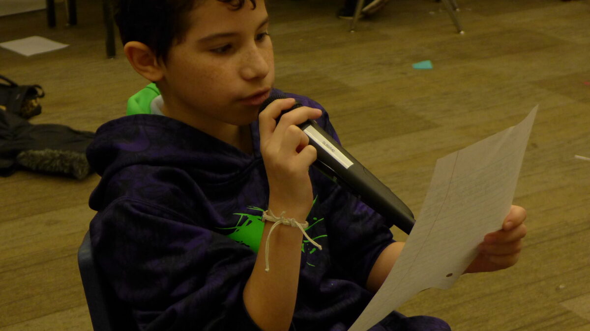 A youth holding a microphone reading a piece of paper in a classroom