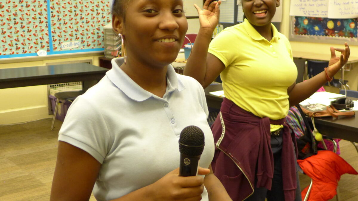 Two youth smiling and standing in a classroom, one holding a microphone