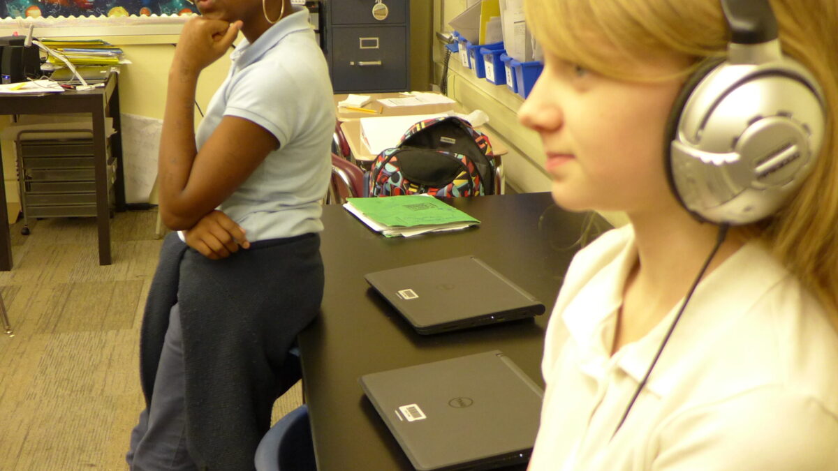 Two youth sitting in front of laptops in a classroom, one wearing headphones