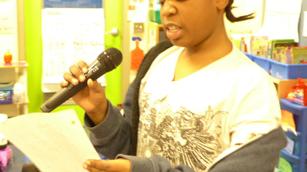 A youth reading a piece of paper and speaking into a microphone in a classroom