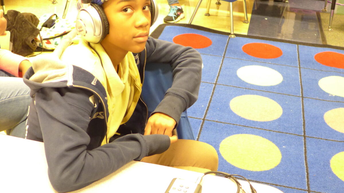 A youth wearing headphones sitting behind a portable recorder on a classroom desk