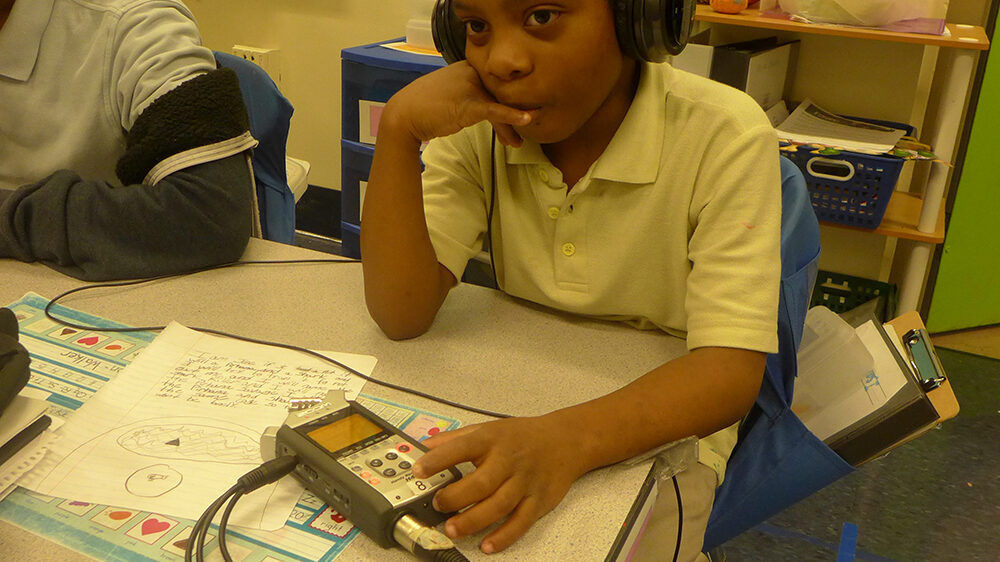 A youth wearing headphones sitting behind a portable recorder on a desk in a classroom