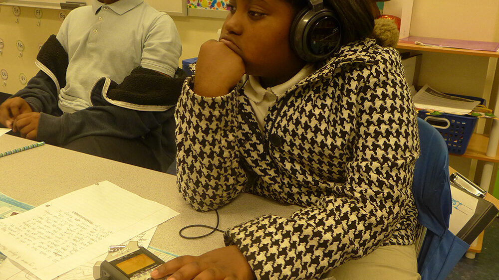 A youth wearing headphones sitting behind a portable recorder on a desk in a classroom.