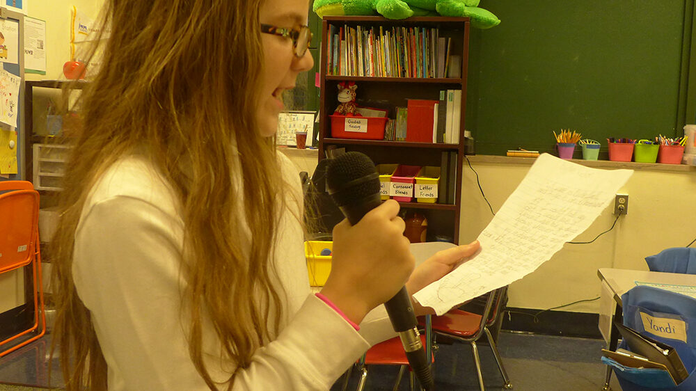 A youth speaking into a microphone and holding a piece of paper in a classroom