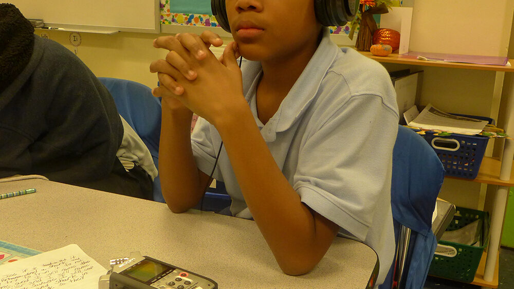 A youth wearing headphones sitting behind a portable recorder on a desk in a classroom
