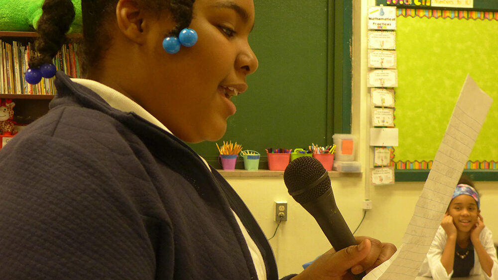A youth reading from a piece of paper and holding a microphone in a classroom