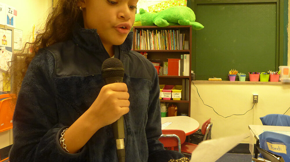 A youth reading from a piece of paper and holding a microphone in a classroom